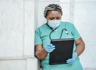 Female nurse in scrubs and mask using tablet for patient care documentation in hospital.