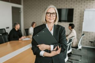 Senior businesswoman with eyeglasses holding a clipboard in a modern conference room with colleagues.